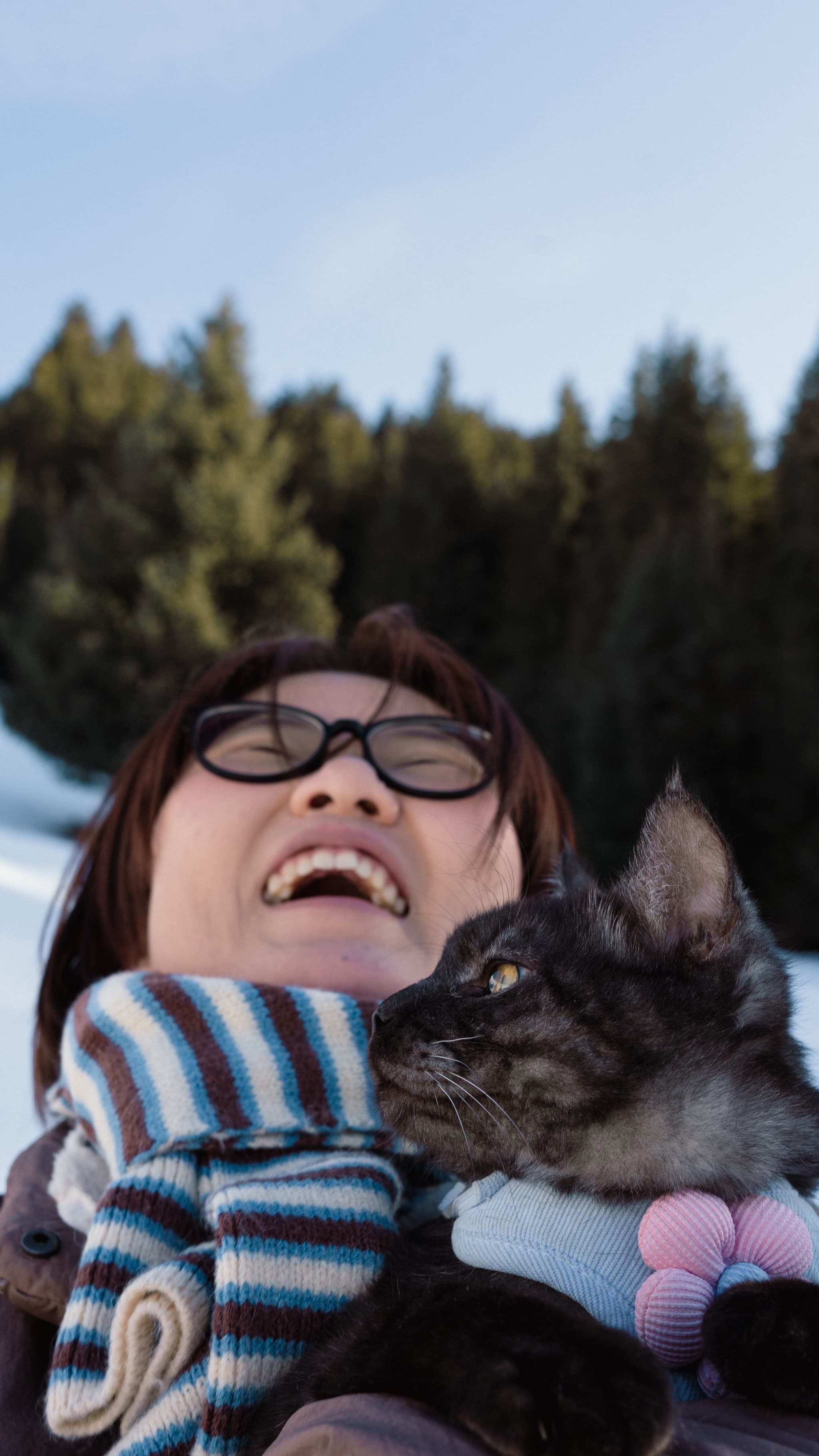 A person wearing glasses and a striped scarf is joyfully holding a cat, with a snowy landscape and trees in the background