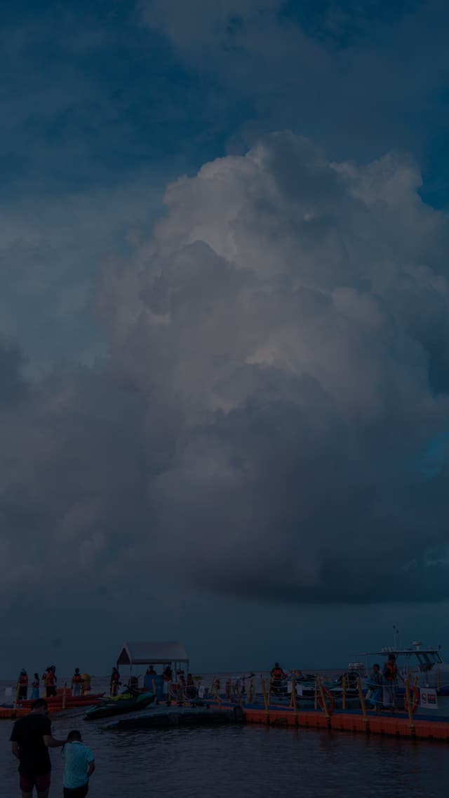 A large, dramatic cloud formation looms over a body of water with people standing on a dock or pier The scene is set during twilight, with a darkening sky