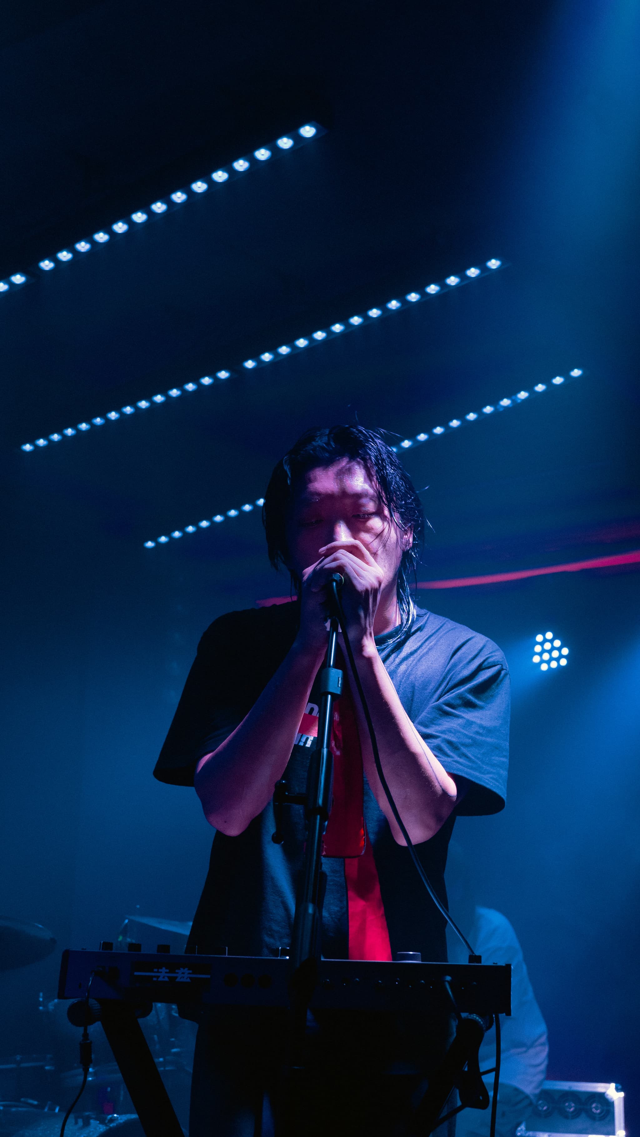 A person singing into a microphone on stage, surrounded by blue and purple lighting, with a keyboard in front