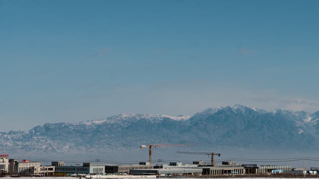 A cityscape with construction cranes in the foreground and a mountain range in the background under a clear blue sky